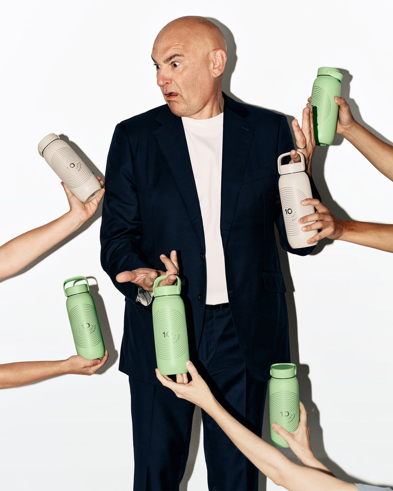 Un homme en costume sombre se tient alors que plusieurs mains présentent des bouteilles d'eau vertes et blanches autour de lui.