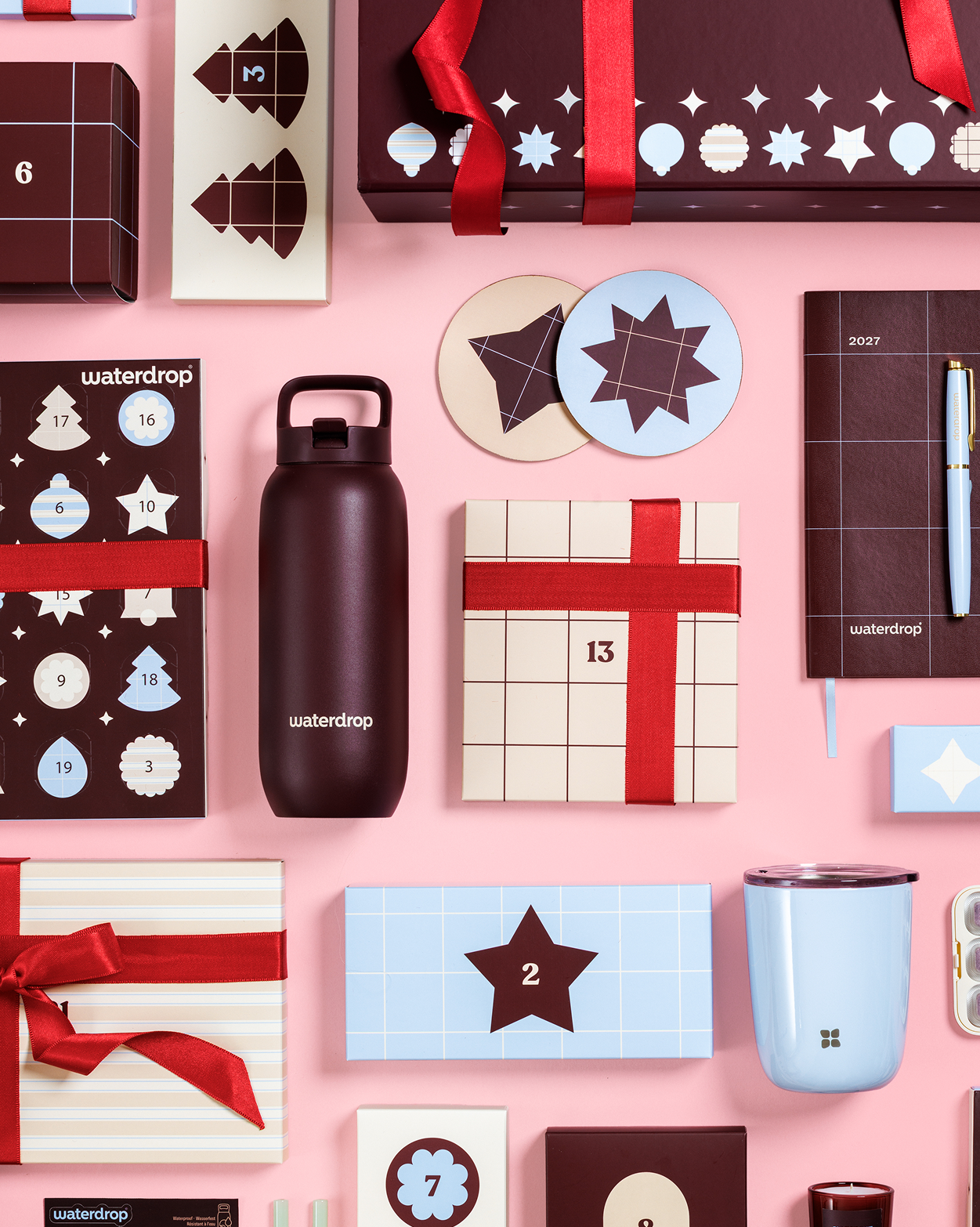 Boîtes-cadeaux variées, calendrier de l'Avent, bouteille, tasse et autres cadeaux emballés sur un fond rose.