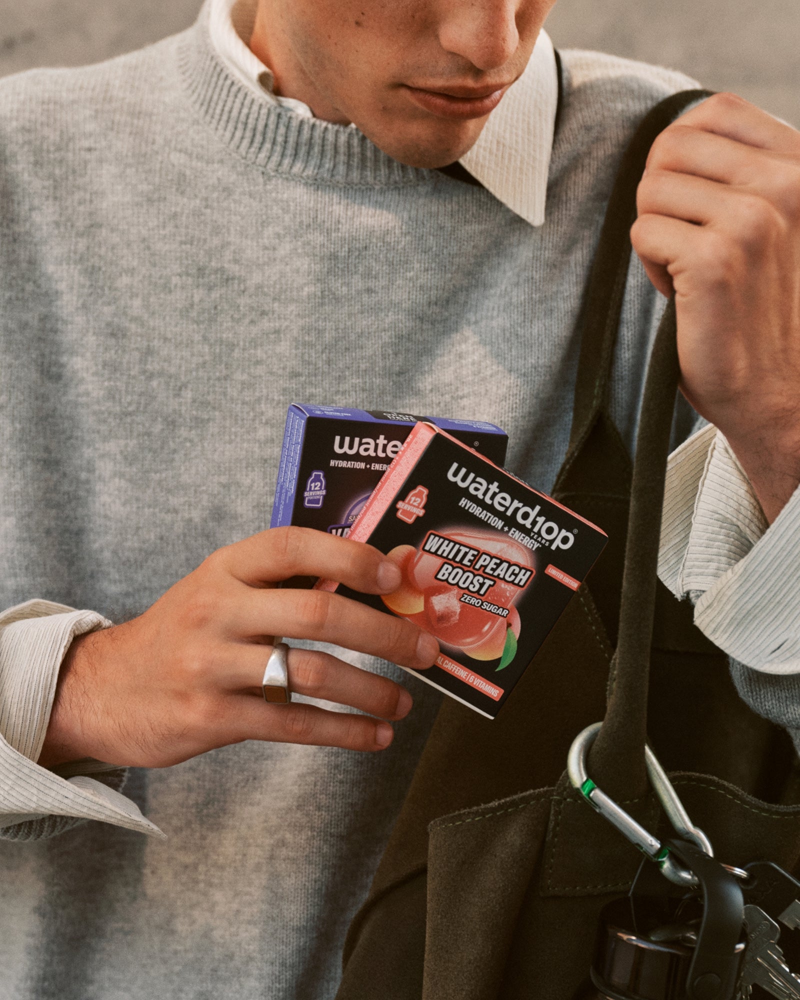 Personne en pull gris tenant de petites boîtes de produit près d'un sac noir.