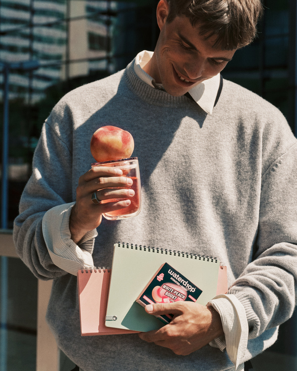 Homme en pull gris dehors tient une boisson rose dans un gobelet avec une pomme sur le bord et un carnet dans l’autre main.