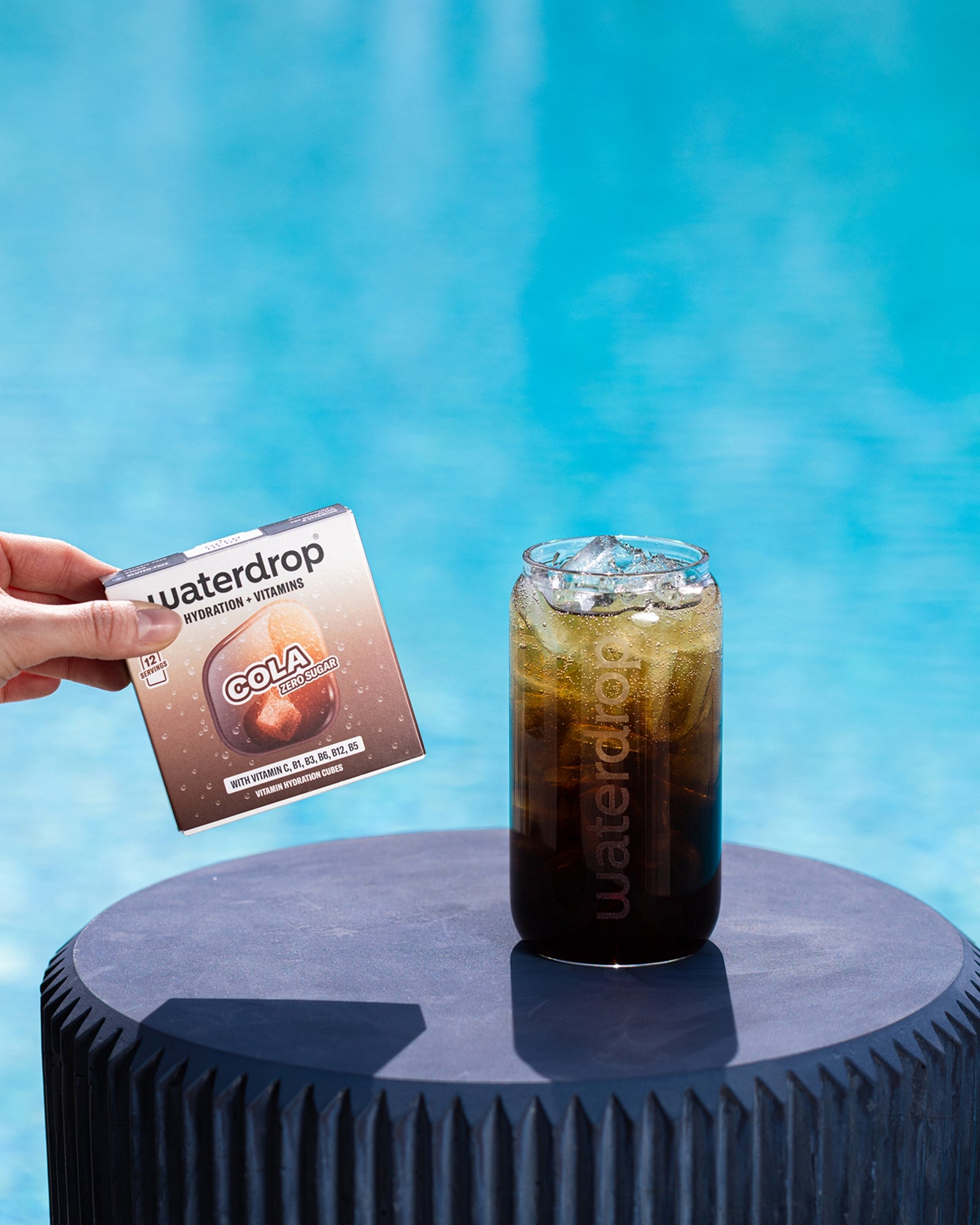 Une main tient un petit sachet à côté d'un verre de cola glacé sur un socle bleu rond près de la piscine.